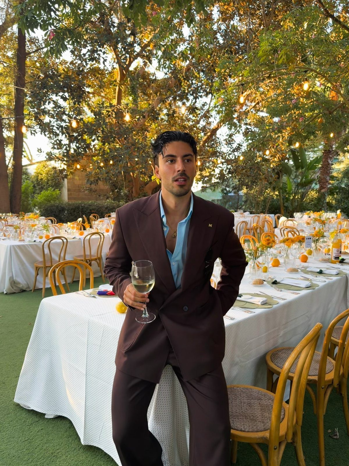 A man wearing a brown suit and blue shirt holding a glass of wine outdoors.