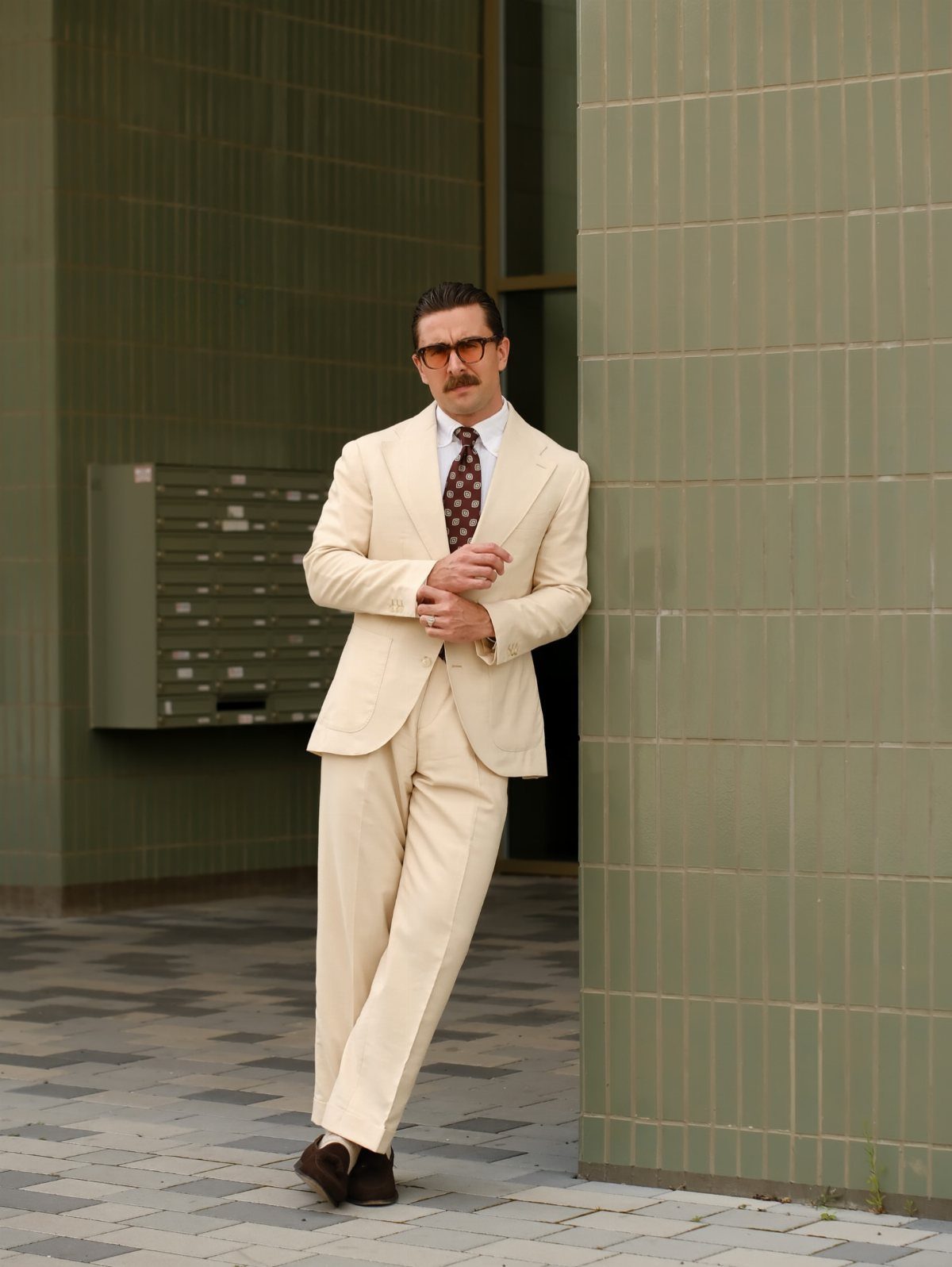 Man in a cream-colored suit and sunglasses leaning against a green tiled wall.
