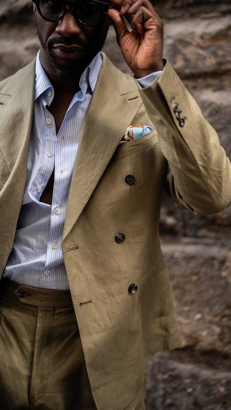 A man in a tan double-breasted suit and striped shirt wearing sunglasses outdoors.