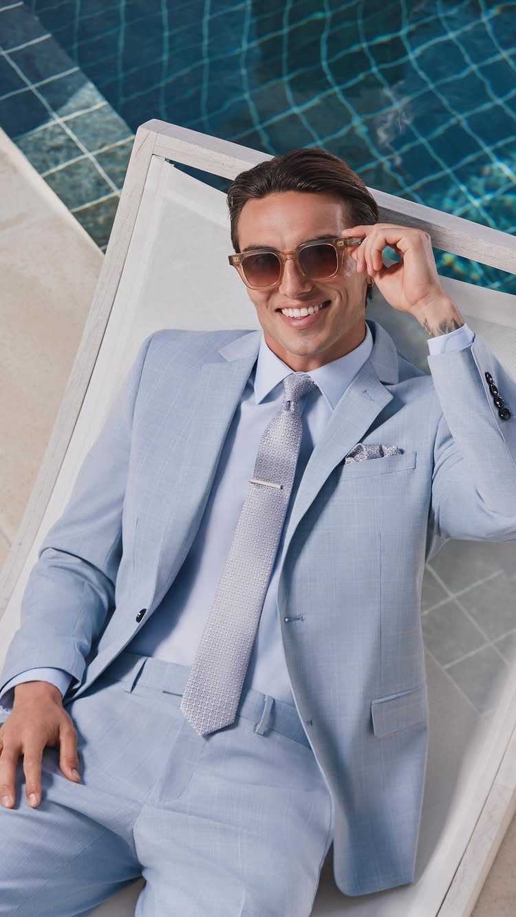 A man in a light blue summer suit and sunglasses lounging by a swimming pool.