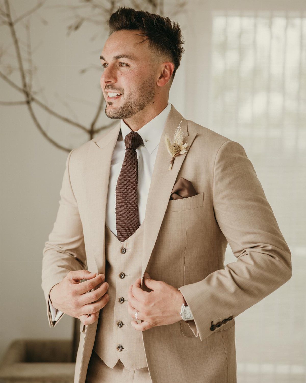Man in a tan three-piece suit with a floral boutonniere and dark tie smiling.