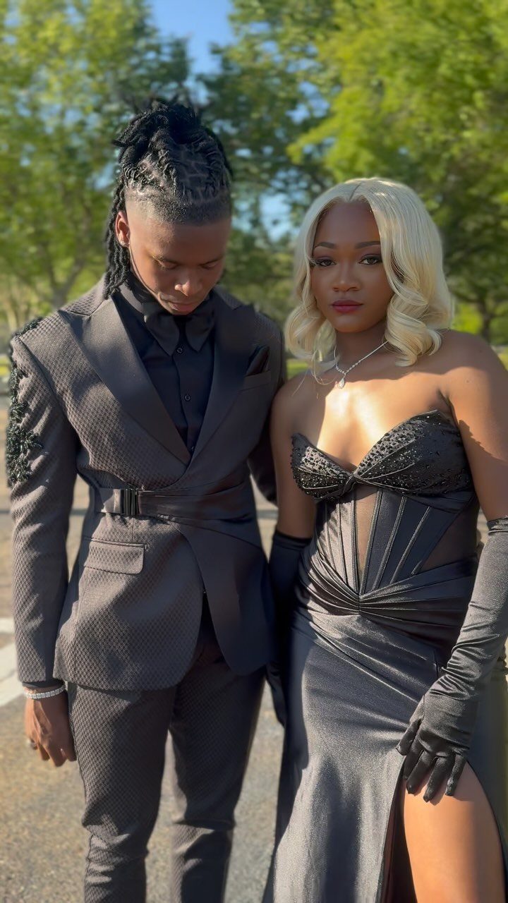 A young man in a black belted suit and a woman in a black gown standing outdoors.