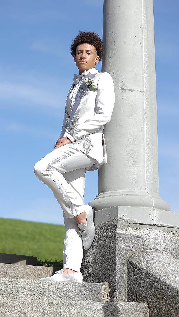 A young man in a white embroidered suit poses against a stone column.