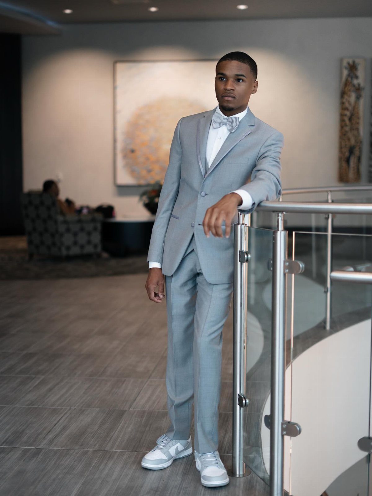 A young man in a light grey suit and patterned bow tie posing by a railing.