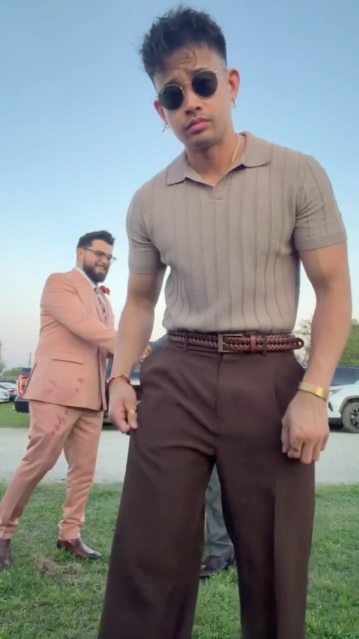 Two men posing outdoors, one in a tan polo and brown trousers, another in a peach suit.