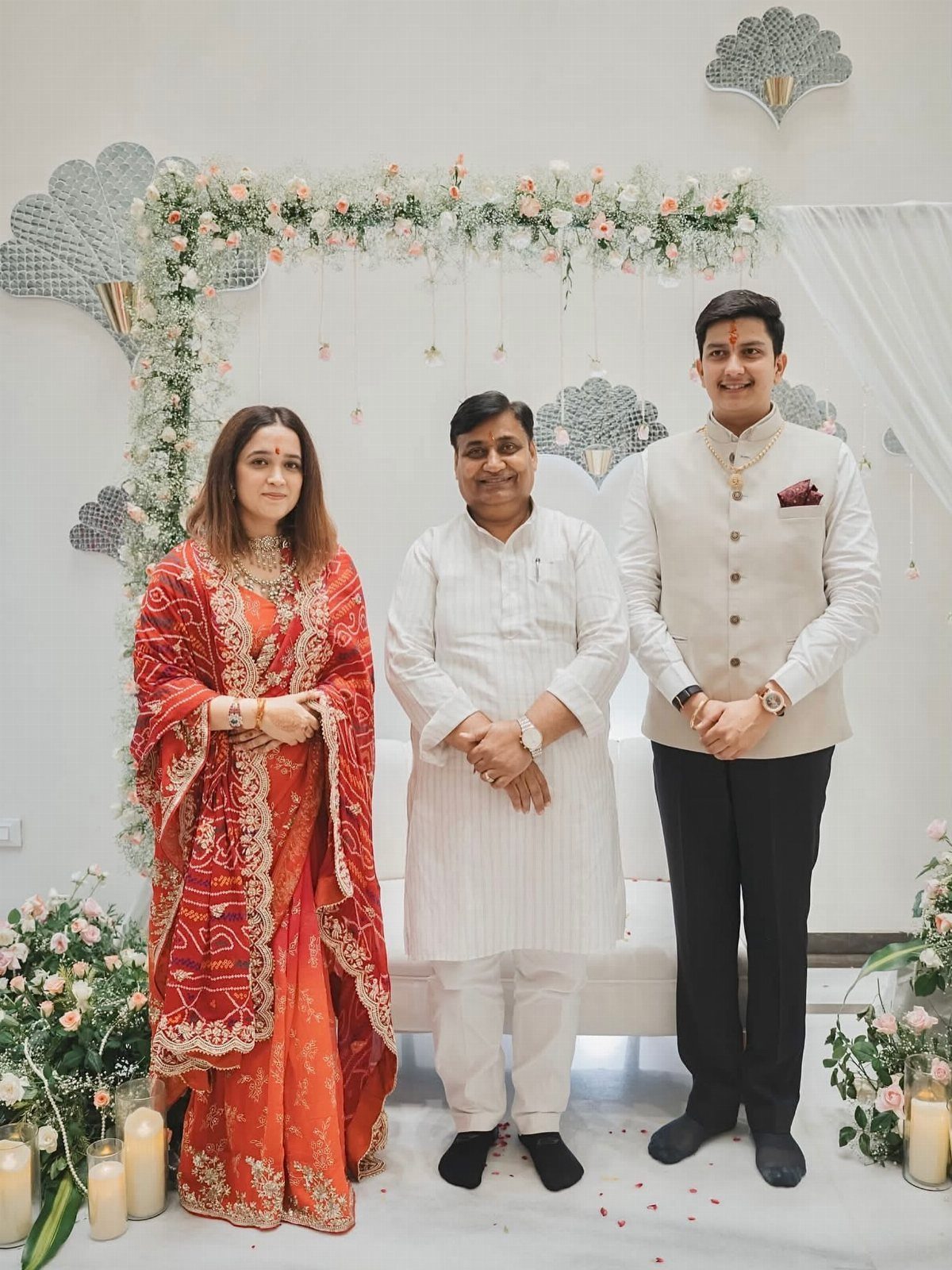 Three people in traditional Indian attire posing in front of a white floral wedding arch.