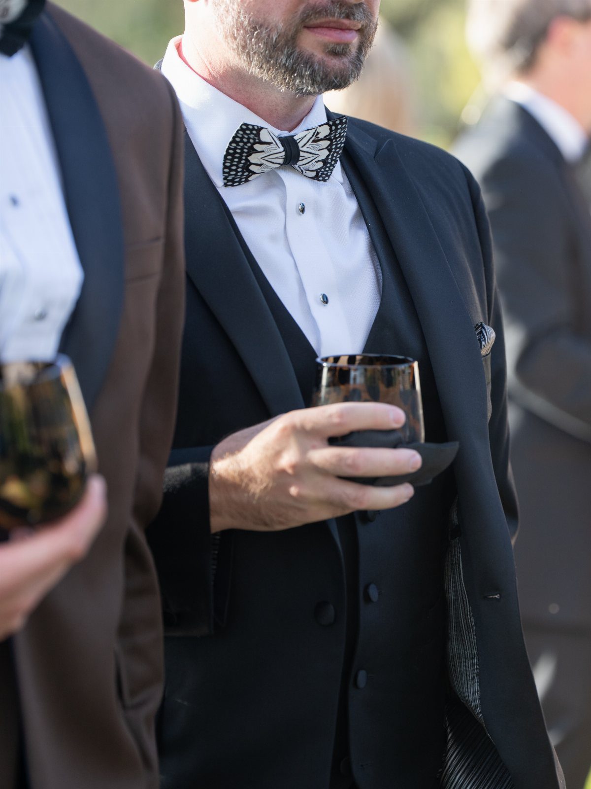 A man wearing a black formal suit and patterned bow tie holding a glass.