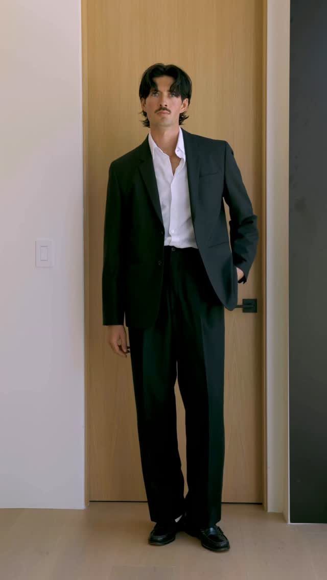 A man posing in a black suit and white shirt against a wooden door.