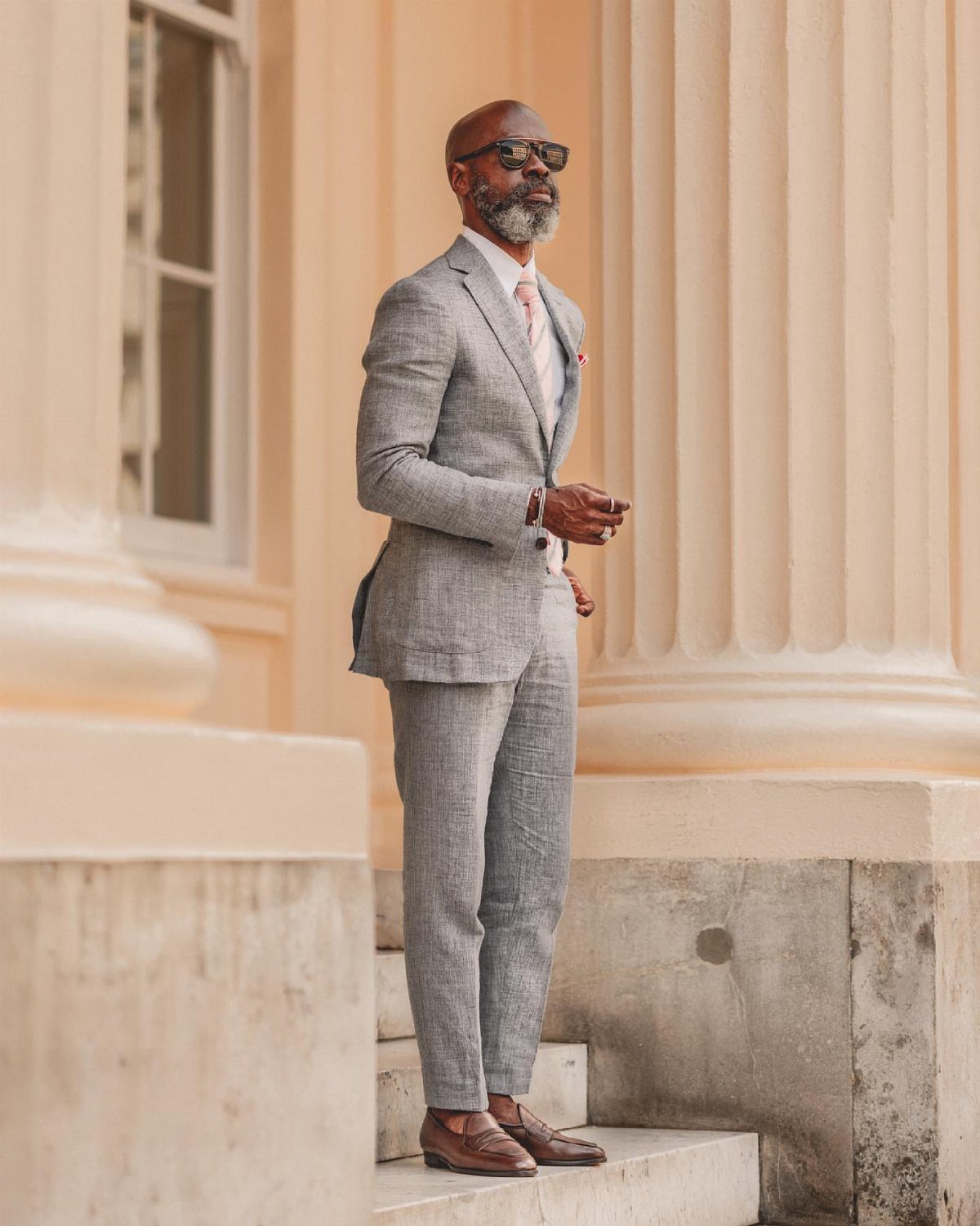 A mature man in a light grey linen suit and pink tie standing on stone steps.