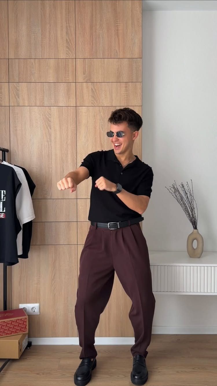 A young man in a black polo and brown trousers posing playfully indoors.