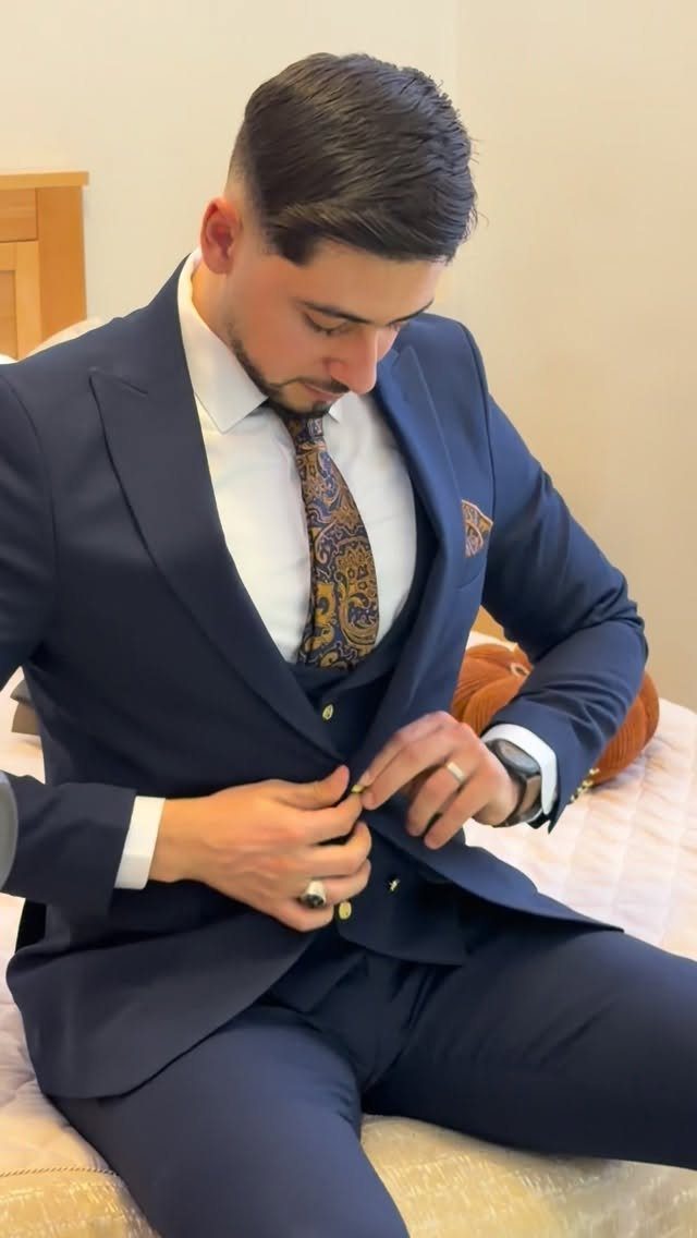 A man in a navy blue three-piece suit buttoning his jacket while sitting on a bed.