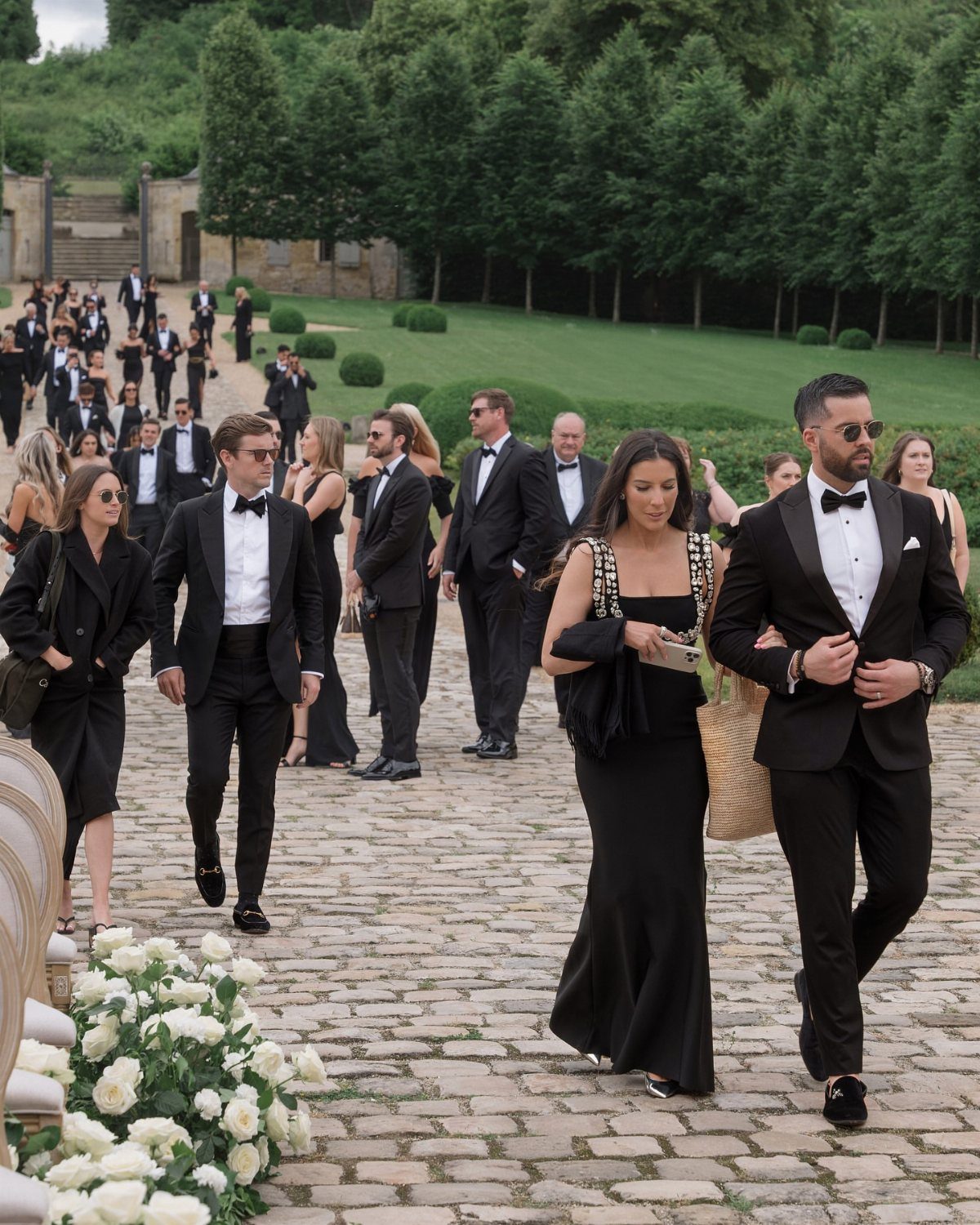 Men in black tuxedos and women in black formal dresses walking on a cobblestone path.