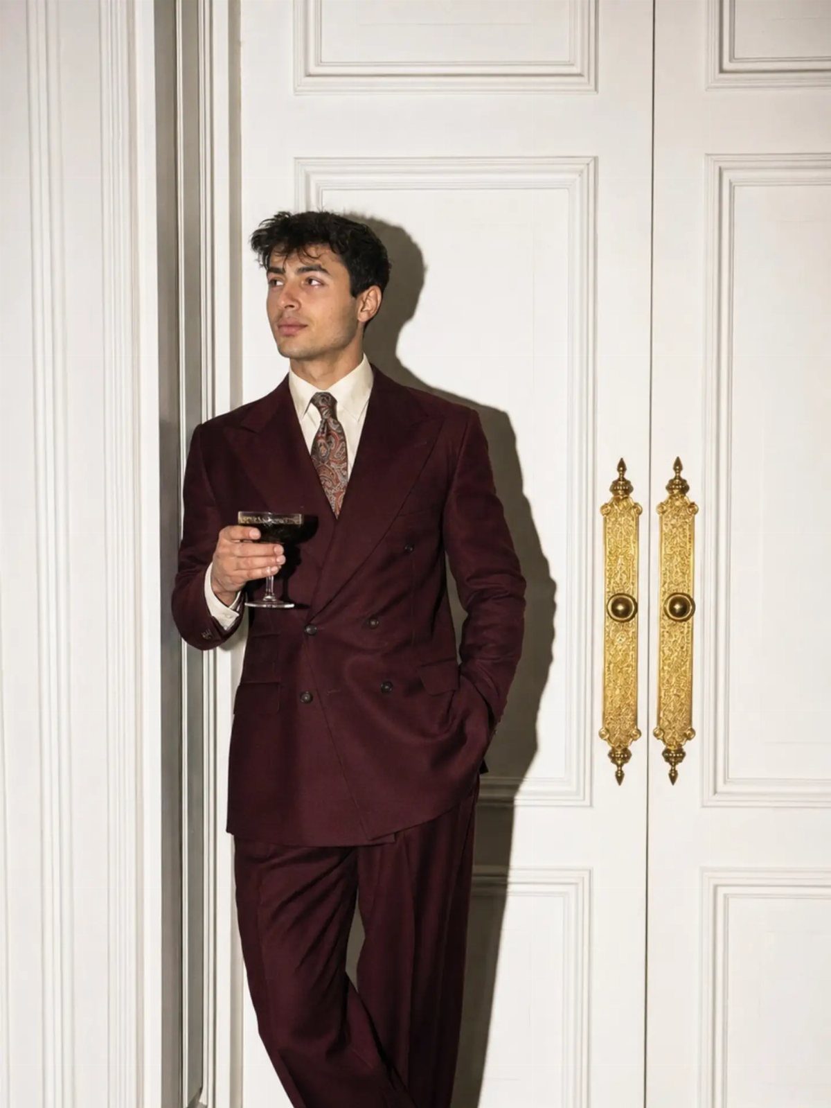 A man in a burgundy double-breasted suit holding a glass against white doors.