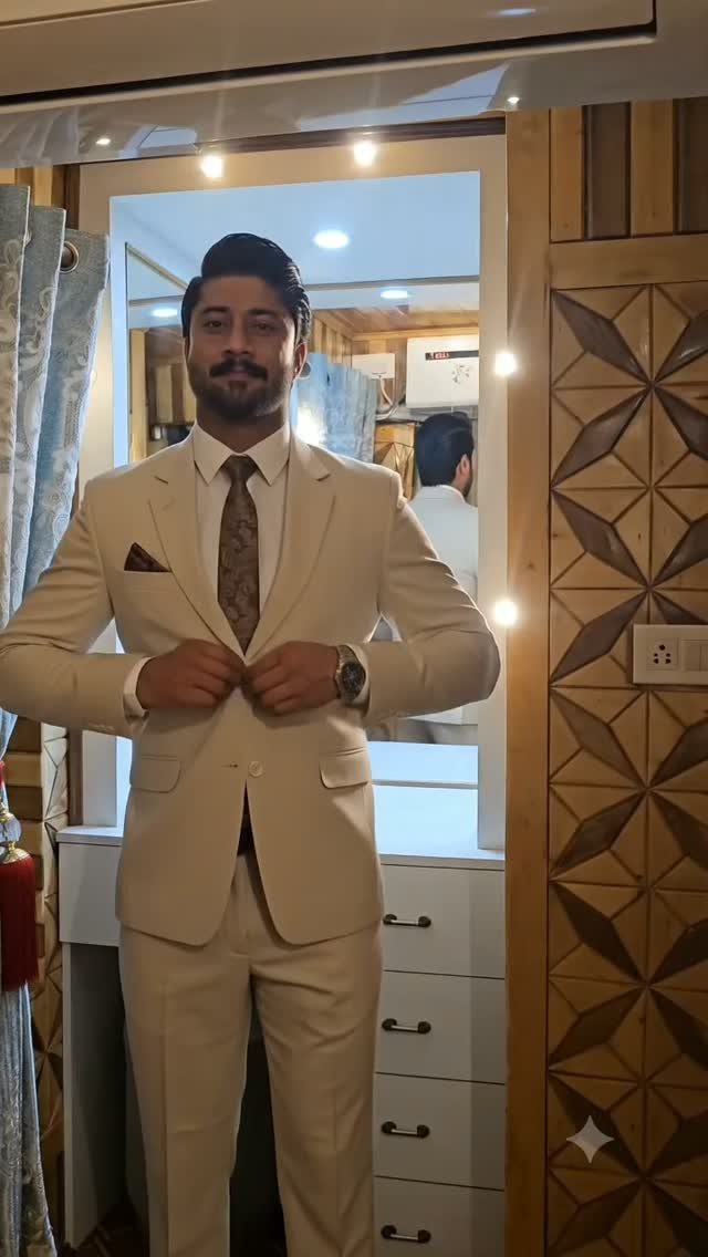 A man smiling while wearing a cream-colored suit and patterned tie in a room.
