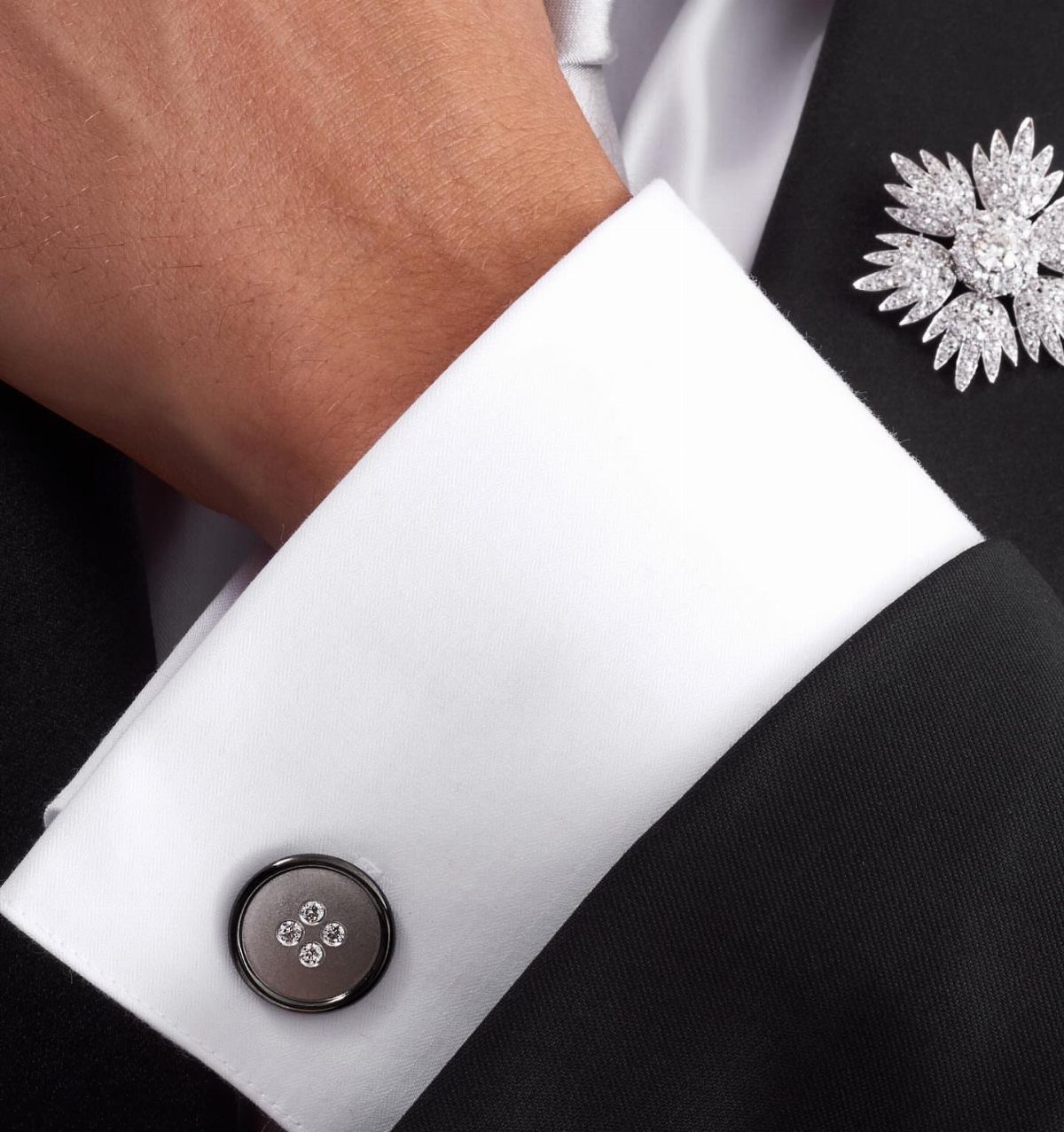 A close-up of a white shirt cuff with a black diamond cufflink and a floral lapel pin.