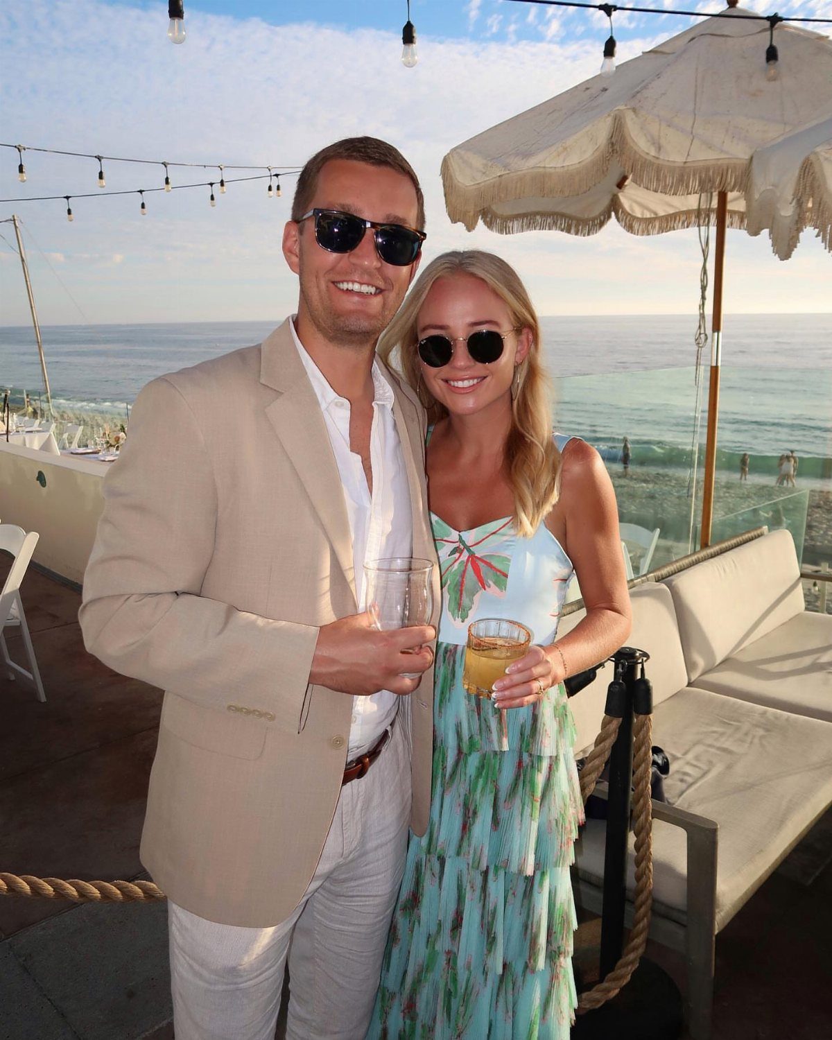 Man in tan blazer and white pants standing with a woman by the ocean.