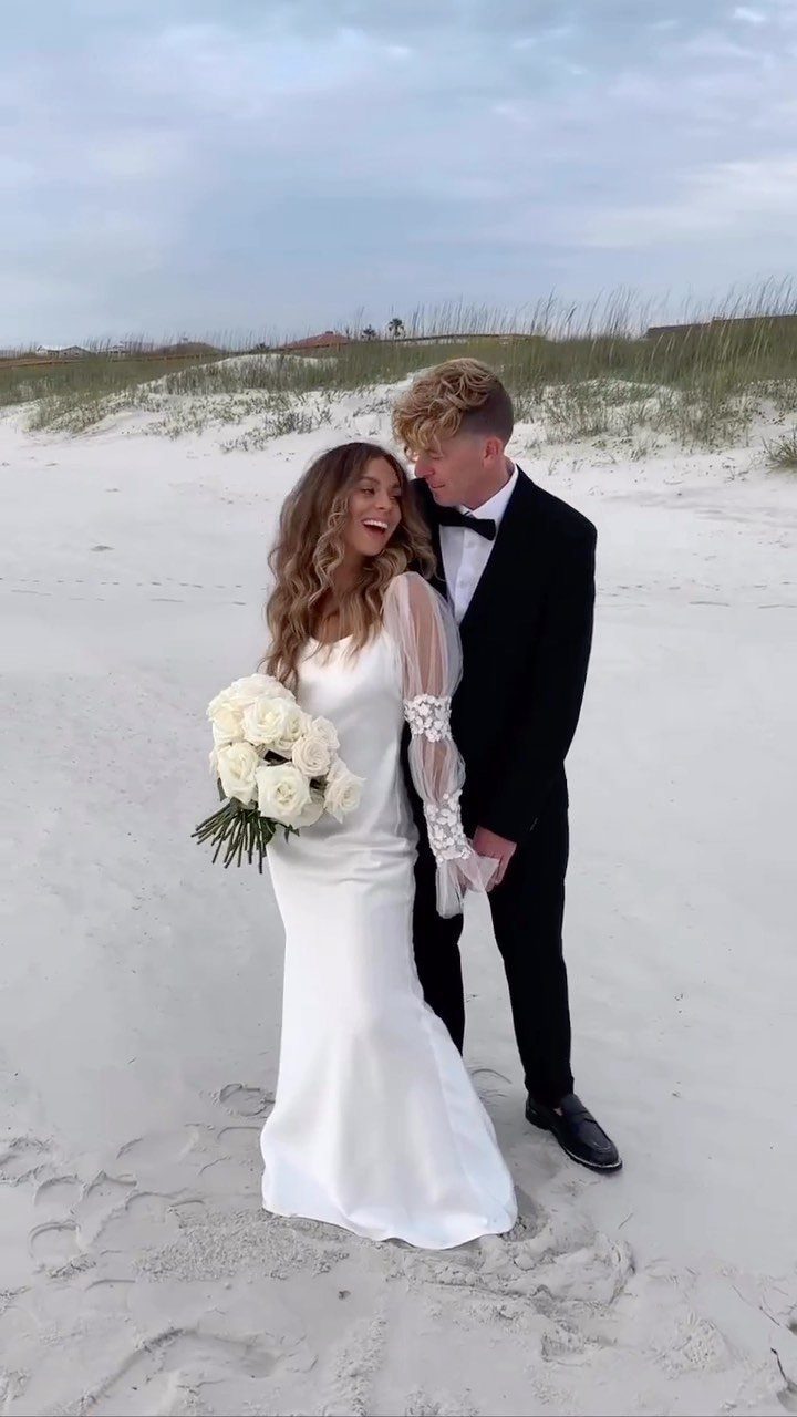 A bride in a white dress and a groom in a black tuxedo standing on a sandy beach.