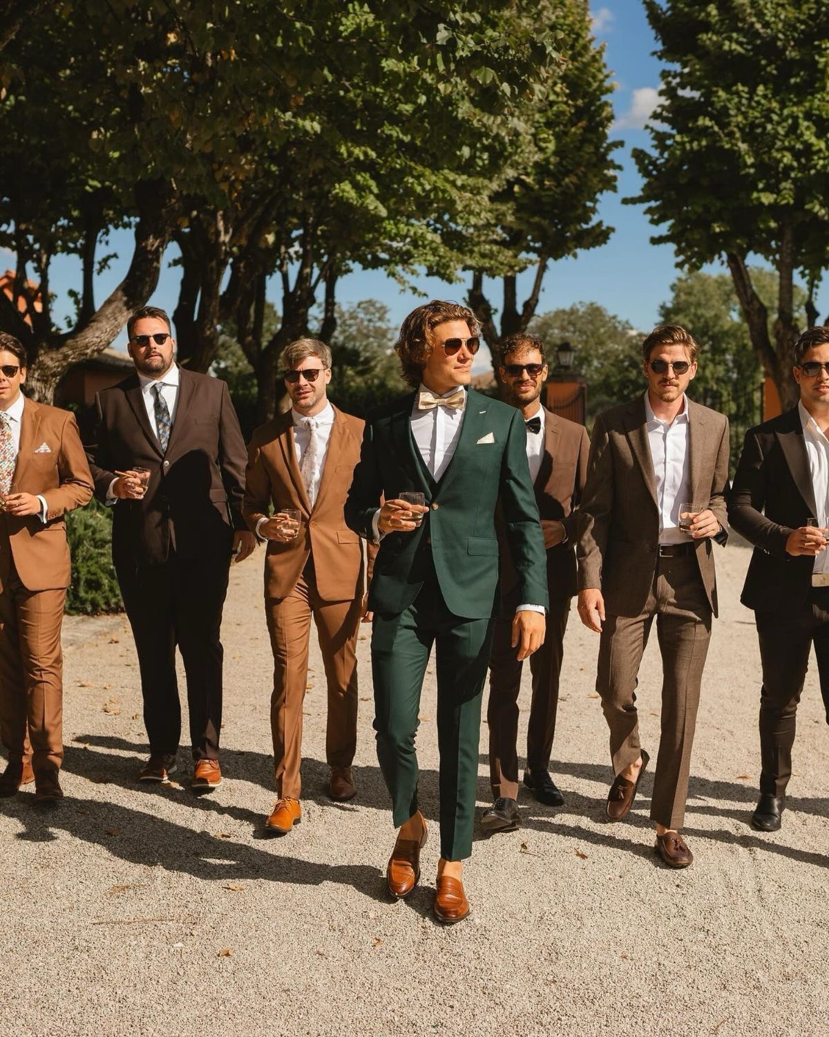 Men in various colored formal suits walking together outdoors on a sunny day.